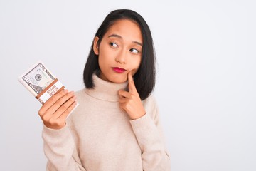 Young beautiful chinese woman holding dollars standing over isolated white background serious face thinking about question, very confused idea