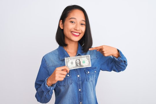 Young beautiful chinese woman holding one dollar standing over isolated white background with surprise face pointing finger to himself