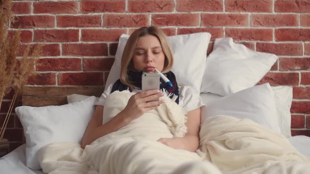 A sick young woman is lying in bed covered with a blanket, texting on the phone and looking at the temperature, taking the thermometer out of her mouth