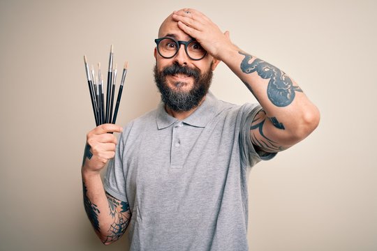 Handsome bald artist man with beard and tattoo painting using painter brushes stressed with hand on head, shocked with shame and surprise face, angry and frustrated. Fear and upset for mistake.