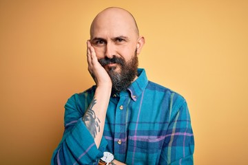 Handsome bald man with beard wearing casual shirt standing over isolated yellow background thinking looking tired and bored with depression problems with crossed arms.