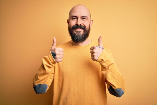 Handsome bald man with beard wearing casual sweater standing over yellow background success sign doing positive gesture with hand, thumbs up smiling and happy. Cheerful expression and winner gesture.