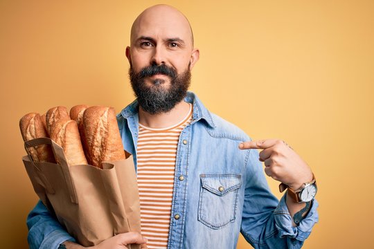 Handsome bald man with beard holding paper bag with bread over yellow background with surprise face pointing finger to himself