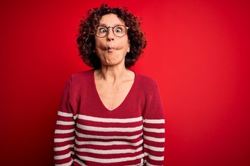 Middle age beautiful curly hair woman wearing casual striped sweater over red background making fish face with lips, crazy and comical gesture. Funny expression.