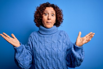 Middle age beautiful curly hair woman wearing casual turtleneck sweater over blue background clueless and confused expression with arms and hands raised. Doubt concept.