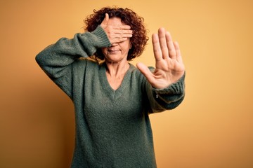 Middle age beautiful curly hair woman wearing casual sweater over isolated yellow background covering eyes with hands and doing stop gesture with sad and fear expression. Embarrassed and negative
