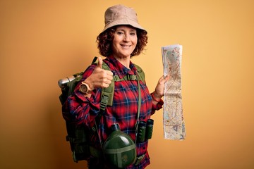 Fototapeta na wymiar Middle age curly hair hiker woman hiking wearing backpack and water canteen holding city map happy with big smile doing ok sign, thumb up with fingers, excellent sign