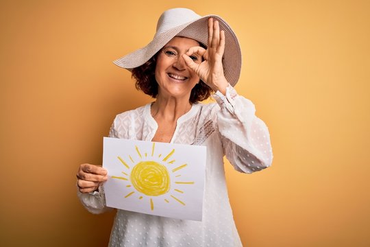 Middle age curly woman on vacation holding bunner with sun image over yellow background with happy face smiling doing ok sign with hand on eye looking through fingers