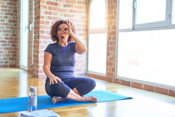 Middle age beautiful sportswoman wearing sportswear sitting on mat practicing yoga at home doing ok gesture shocked with surprised face, eye looking through fingers. Unbelieving expression.