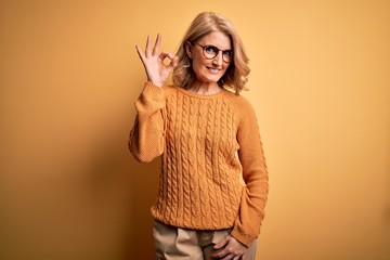 Middle age beautiful blonde woman wearing casual sweater and glasses over yellow background smiling...