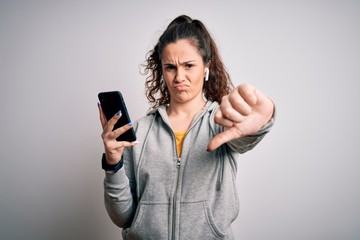 Naklejka premium Beautiful sportswoman with curly hair listening to music using smartphone and earphones with angry face, negative sign showing dislike with thumbs down, rejection concept