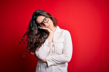 Young beautiful woman with curly hair wearing shirt and glasses over red background thinking looking tired and bored with depression problems with crossed arms.