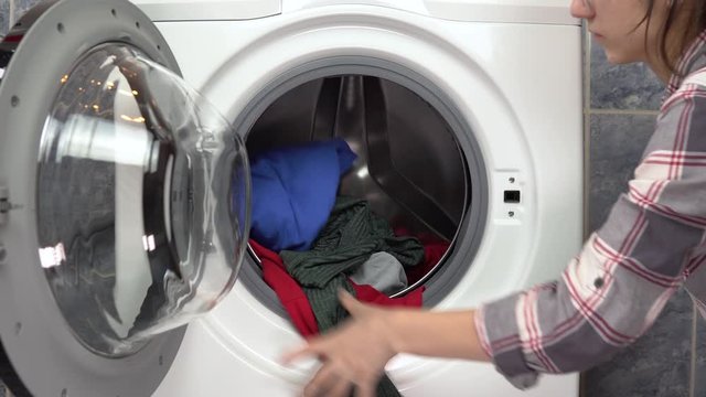 A young woman is actively loading clothes with a washing machine. The woman opens the washing machine and throws clothes into her then closes it.
