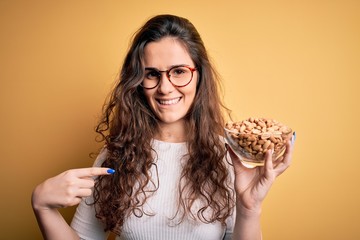Beautiful woman with curly hair holding bowl with healthy peanuts over yellow background with surprise face pointing finger to himself