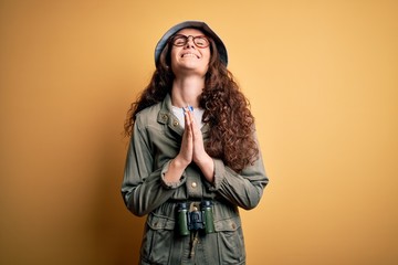 Young beautiful tourist woman on vacation wearing explorer hat and binoculars begging and praying...