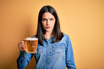 Young woman with blue eyes drinking jar of beer standing over isolated yellow background skeptic...