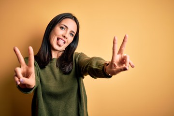 Young brunette woman with blue eyes wearing green casual sweater over yellow background smiling with tongue out showing fingers of both hands doing victory sign. Number two.
