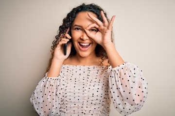 Young beautiful woman with curly hair having conversation talking on the smartphone with happy face smiling doing ok sign with hand on eye looking through fingers