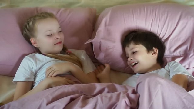 Funny adorable happy kids girl and boy playing on bed together. cute little brother sister children in white pajamas having fun in bedroom. preteen laughing and enjoying morning.  quarantine vacation