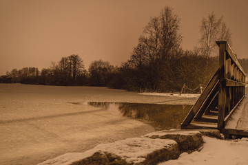 Drweniany mały mostek na zamarźniętym jeziorze w mroźny zimowy wieczór © Dreamnordno