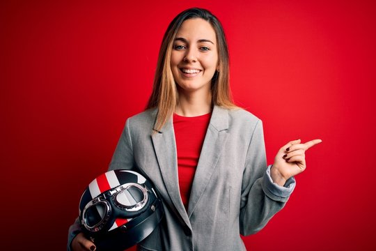Young beautiful blonde motorcyclist woman holding motorcycle helmet over red background very happy pointing with hand and finger to the side
