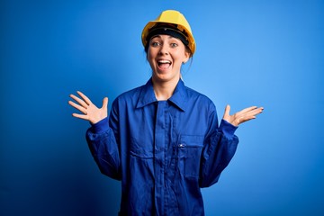 Young beautiful worker woman with blue eyes wearing security helmet and uniform celebrating crazy and amazed for success with arms raised and open eyes screaming excited. Winner concept