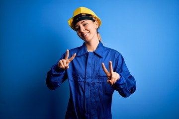 Young beautiful worker woman with blue eyes wearing security helmet and uniform smiling looking to the camera showing fingers doing victory sign. Number two.