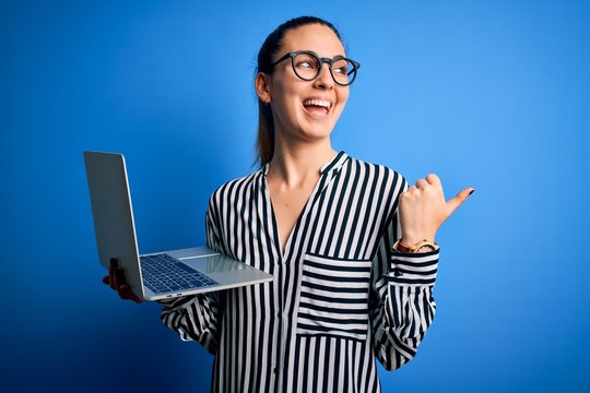 Young beautiful blonde businesswoman wearing glasses using laptop over blue background pointing and showing with thumb up to the side with happy face smiling