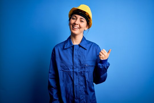 Young beautiful worker woman with blue eyes wearing security helmet and uniform smiling with happy face looking and pointing to the side with thumb up.