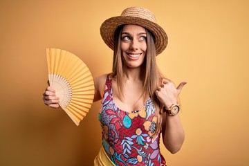 Young beautiful blonde woman wearing swimsuit and summer hat holding fan over yellow background pointing and showing with thumb up to the side with happy face smiling