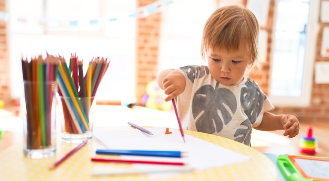 Adorable toddler drawing using paper and pencil around lots of toys at kindergarten