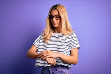 Young beautiful blonde woman wearing striped t-shirt and glasses over purple background Checking the time on wrist watch, relaxed and confident