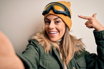 Young brunette skier woman wearing snow clothes and ski goggles make selfie by camera very happy pointing with hand and finger
