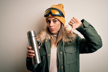 Young beautiful brunette skier woman wearing ski goggles drinking thermo with coffe with angry...