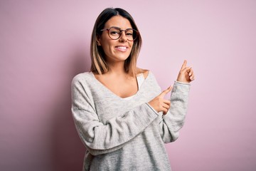 Young beautiful brunette woman wearing casual sweater and glasses over pink background smiling and looking at the camera pointing with two hands and fingers to the side.