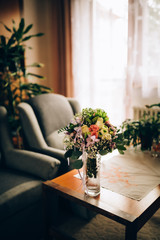 Bouquet composed of roses, other types of flowers, decorative leaves and completed with a white ribbon with lace. The bouquet is placed in a glass vase,which stands on an old living room coffee table.