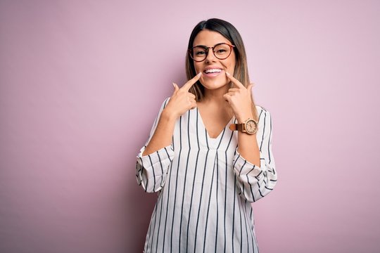 Young beautiful woman wearing casual striped t-shirt and glasses over pink background Smiling with open mouth, fingers pointing and forcing cheerful smile