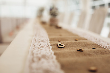 Detail of decorated dining table with burlap tablecloth and white decorative lace. On tablecloth are placed wooden ornaments in shape of small hearts. Shallow depth of field. Background out of focus.