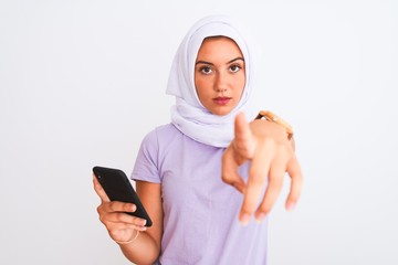 Young beautiful arabian girl wearing hijab using smartphone over isolated white background pointing with finger to the camera and to you, hand sign, positive and confident gesture from the front