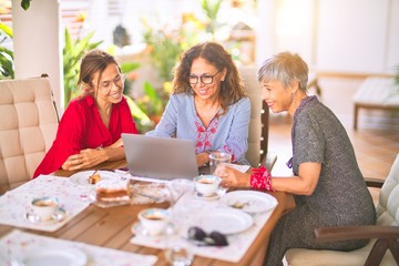 Meeting of middle age women having lunch and drinking coffee. Mature friends smiling happy using laptop at home on a sunny day