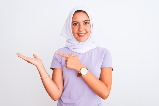 Young beautiful arabian girl wearing hijab standing over isolated white background amazed and smiling to the camera while presenting with hand and pointing with finger.