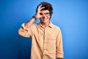 Young handsome man wearing casual shirt standing over isolated blue background doing ok gesture with hand smiling, eye looking through fingers with happy face.