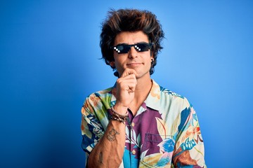 Young handsome man wearing thug life glasses standing over isolated blue background with hand on chin thinking about question, pensive expression. Smiling with thoughtful face. Doubt concept.