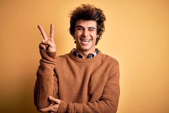 Young handsome man wearing casual shirt and sweater over isolated yellow background smiling with happy face winking at the camera doing victory sign with fingers. Number two.