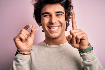 Young handsome man holding aligner standing over isolated pink background surprised with an idea or question pointing finger with happy face, number one
