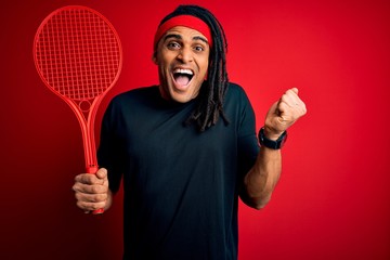 African american afro sportsman with dreadlocks holding tennis racket over red background screaming proud and celebrating victory and success very excited, cheering emotion