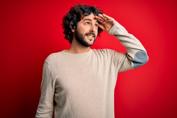 Fototapeta na wymiar Young handsome man with beard wearing casual sweater standing over red background very happy and smiling looking far away with hand over head. Searching concept.