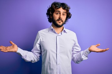 Young handsome business man with beard wearing shirt standing over purple background clueless and confused expression with arms and hands raised. Doubt concept.