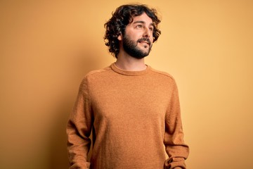 Young handsome man with beard wearing casual sweater standing over yellow background looking away to side with smile on face, natural expression. Laughing confident.