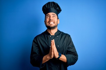 Young handsome chef man with beard wearing cooker uniform and hat over blue background begging and praying with hands together with hope expression on face very emotional and worried. Begging.